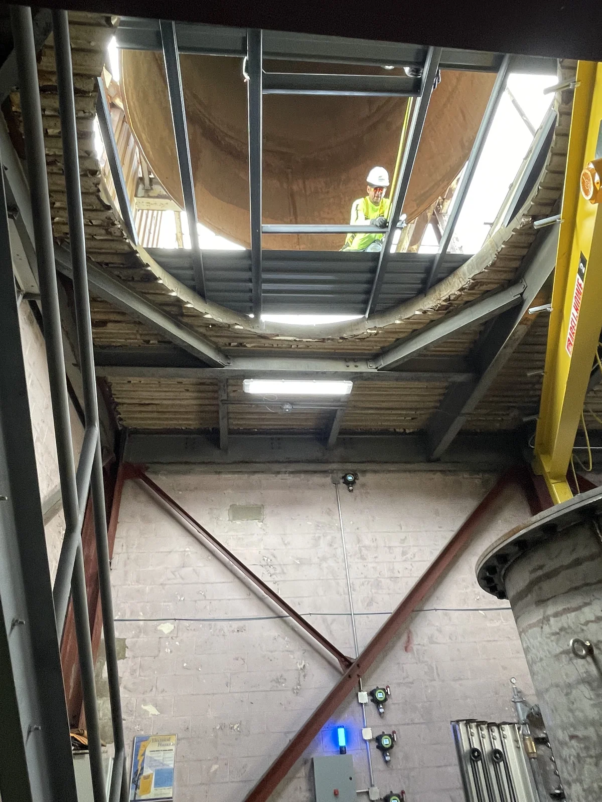Looking up through a steel-framed roof opening inside a brick industrial building at a worker in a hard hat beneath a rooftop tank